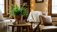 Cozy living room corner with fern plant, wooden table, leather armchair, knit throw, and warm fairy lights creating a hygge atmosphere.