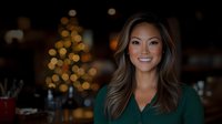 Young Asian woman with radiant smile wearing green blouse against blurred Christmas lights and festive bokeh background, creating warm holiday atmosphere.