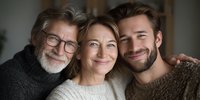 Multi-generational family portrait showing senior man with glasses, middle-aged woman, and young bearded man smiling warmly together in cozy sweaters.