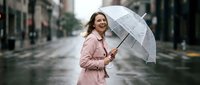 Young woman with long brown hair and fair complexion wearing a pink jacket, holding a transparent umbrella on a wet urban street during rainfall. Suitable for weather, lifestyle, and seasonal campaigns.