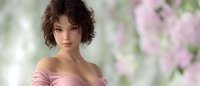 Young Asian woman with curly dark hair in pink off-shoulder dress against soft floral background with cherry blossoms, romantic spring portrait with gentle natural lighting.