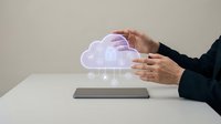 Businessman hands protecting holographic cloud computing icon above tablet on white desk representing data security and digital technology solutions.