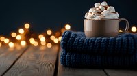 Cozy hot chocolate topped with marshmallows on knitted blue scarf, wooden table with blurred golden bokeh lights creating warm winter atmosphere.