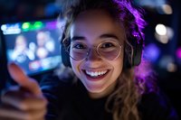 Young Asian female gamer wearing headphones and glasses smiles brightly in purple neon lighting while streaming or playing video games, showing thumbs up gesture.