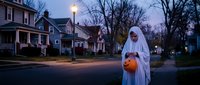 Child in white ghost costume holding orange pumpkin bucket trick-or-treating on suburban street at dusk during Halloween celebration.