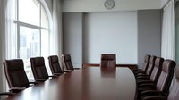 Modern corporate boardroom with large wooden conference table and leather executive chairs overlooking city skyline through floor-to-ceiling windows.