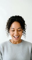 Young woman with dark curly hair, medium brown skin tone, wearing light gray sweater, smiling with eyes closed against white background for portrait and lifestyle use.