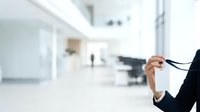 Businesswoman in formal black attire holding eyeglasses in modern office corridor with blurred background, suitable for corporate, professional, and business lifestyle content.
