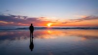 Silhouetted surfer holding board on wet beach at golden hour sunset, reflecting in calm water, ideal for travel and lifestyle content.