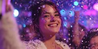 Young woman smiling at nightclub with colorful purple lighting and falling confetti, enjoying festive atmosphere in fluffy white outfit.