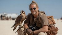Young Caucasian man with falcon on sandy beach, wearing sunglasses and casual clothing with camera and leather bag, enjoying wildlife photography adventure.