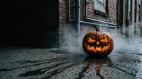 Carved jack-o-lantern with glowing face sits on wet pavement in front of old brick building with fog, creating spooky atmosphere for seasonal decorations and events.