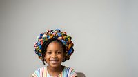 Young girl with African descent wearing colorful traditional head wrap, smiling at camera against neutral background. Suitable for diversity, cultural representation, education, and family-themed designs.