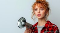 Young Caucasian woman with curly red hair holding megaphone wearing red plaid shirt against gray background for communication and announcement concepts.
