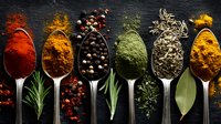 Colorful spices and herbs arranged in silver spoons on dark background, including paprika, turmeric, peppercorns, herbs, and curry powder for cooking and seasoning.