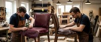Two skilled craftsmen restoring vintage burgundy leather chair in professional furniture workshop with tools and wooden frames in background.