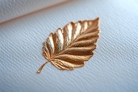Golden metallic leaf brooch with detailed veining pattern on textured white background, elegant jewelry accessory for fashion and design projects.