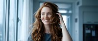 Young professional woman with curly red hair smiling confidently in modern office environment. Business portrait for corporate communications and workplace diversity.