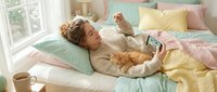Young woman relaxing in bed with orange tabby cat while using smartphone in cozy pastel bedroom with colorful pillows and morning coffee cup nearby.