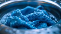 Vibrant blue fabric submerged in washing machine water with bubbles and soap suds, creating dynamic texture and ripples in macro photography view.