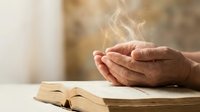 Elderly hands cupped in prayer position above open book with warm light streaming through creating peaceful spiritual atmosphere for meditation and faith.