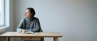 Young Asian woman in gray sweater sitting at wooden desk by window, looking thoughtful and contemplative in minimalist modern interior space.