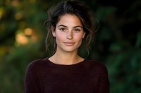 Young Mediterranean woman with natural makeup and messy updo hairstyle wearing burgundy sweater smiling at camera against blurred green outdoor background.