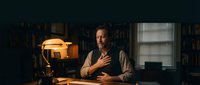 Middle-aged Caucasian man sitting thoughtfully at wooden desk in dimly lit library office with warm lamp light, books on shelves, contemplative professional atmosphere.