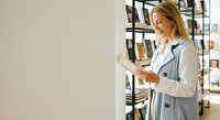 Young Caucasian woman with blonde hair wearing a light blue blazer and white shirt, reading a book in a modern library or bookstore with shelves in the background, suitable for education, learning, and lifestyle content.