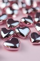 Metallic rose gold heart shapes scattered on pink surface with reflective chrome finish, creating romantic Valentine's Day or wedding design background.