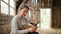 Woman with brown goat in rustic barn setting, wearing beige sweater, holding young animal with care and affection during farm work.