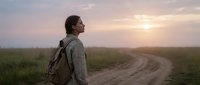 Male traveler with backpack standing on rural dirt road at sunrise, gazing toward horizon across misty green fields. Adventure and exploration concept.