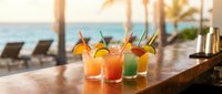 Colorful tropical cocktails with fruit garnishes on wooden bar overlooking ocean beach resort with palm trees and lounge chairs during golden hour sunset.