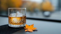 Crystal whiskey glass with amber liquid and ice cube on dark surface with autumn maple leaf, warm bokeh background for luxury beverage advertising.