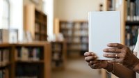 Person of African descent holding open blank book in library setting with shelves and natural light in background.