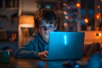 Young Caucasian boy illuminated by laptop screen in dark room with bokeh lights, depicting late night internet usage, digital addiction, or cybersecurity concerns.