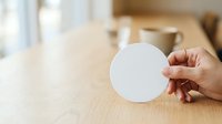 Close-up of hand holding blank white circular card on wooden table with blurred office background, suitable for branding mockups and corporate presentations.