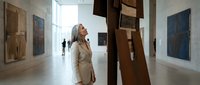 Woman with long gray hair wearing beige blazer viewing large wooden cross sculpture in contemporary art gallery with natural light and minimalist interior design.