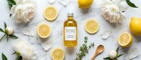 Overhead flat lay of golden oil bottle surrounded by fresh lemon slices, white peonies, herbs and wooden spoon on marble surface for culinary and wellness design projects.