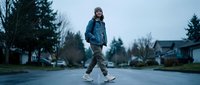 Young male child, Caucasian ethnicity, wearing blue jacket and winter beanie, jogging on suburban residential street with evergreen trees and houses in background during overcast day.