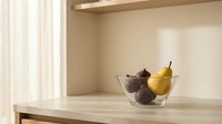 Glass bowl with fresh pears and figs on wooden table in bright, minimalist kitchen interior with natural light from window.