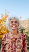 Mature woman with silver hair wearing floral boho dress smiles genuinely outdoors against autumn foliage background in natural sunlight.