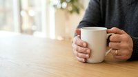 Person with medium skin tone holding white ceramic mug at wooden table, wearing dark sweater, relaxed morning beverage moment for lifestyle and wellness content.