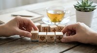 Hands arranging wooden blocks with wellness icons on rustic table with herbal tea and succulent plant for mindful living and self-care concepts.