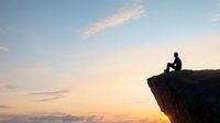 Silhouette of person sitting alone on cliff edge against dramatic sunset sky, contemplating nature and life, peaceful solitary moment in wilderness landscape.