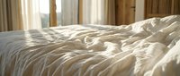 Unmade bed with cream-colored linens and pillows in bright morning light from windows with beige curtains, conveying comfort and relaxation for bedroom design projects.