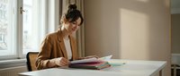 Young Asian businesswoman in brown blazer reading documents at modern office desk by window with natural lighting for professional workplace concepts.
