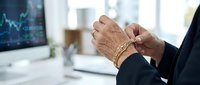 Mature male professional wearing dark business suit and gold bracelet gestures with hands during financial discussion in modern office with stock market data on screen.