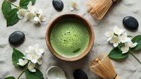 Matcha green tea in wooden bowl with bamboo whisk, jasmine flowers, and black stones on linen background, top view flat lay.