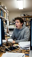 Young Caucasian male student with curly brown hair wearing gray hoodie, expressing surprise while working at computer desk in library office surrounded by books, shelves, and study materials.